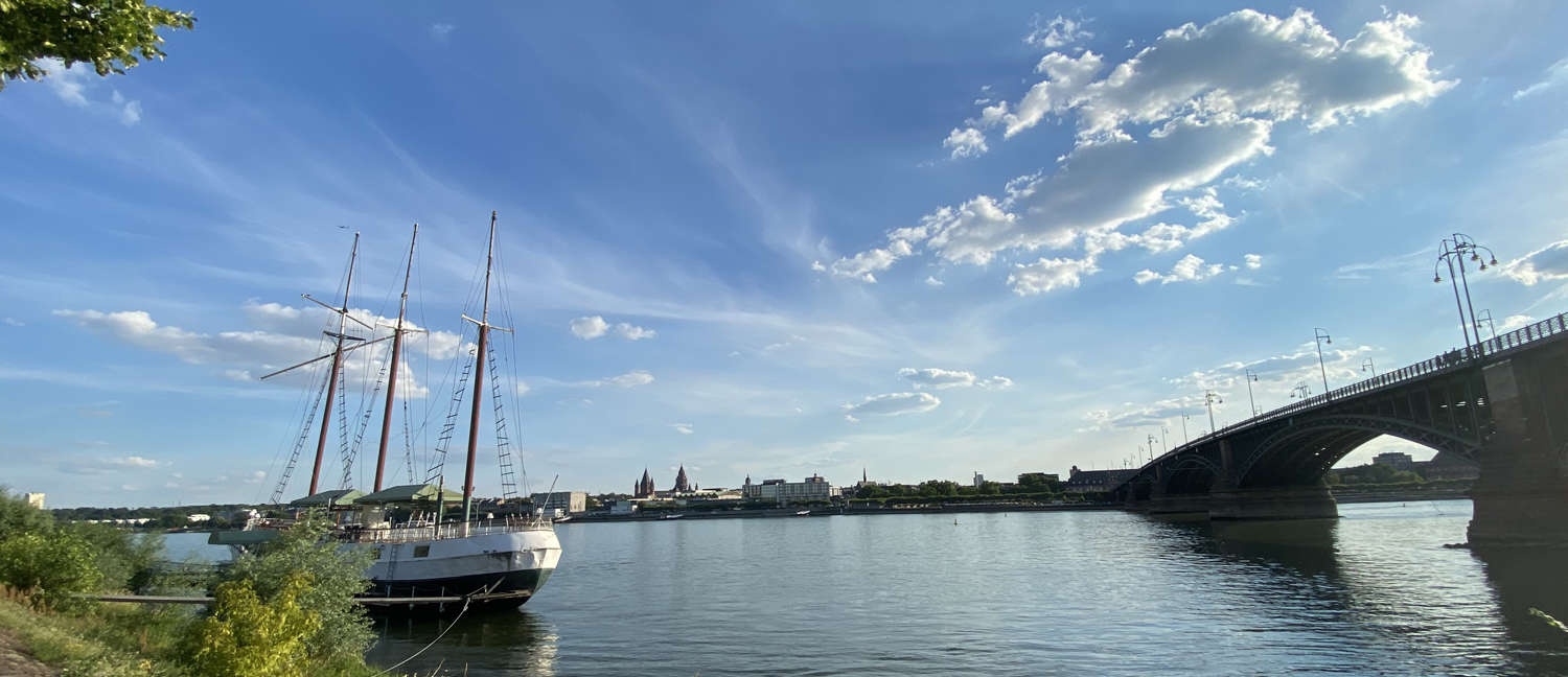 Blick auf Mainz Segelschiff mit eingeholten Segeln links auf Fluss. Rechts eine Brücke. Im Hintergrund Stadtsilhouette.