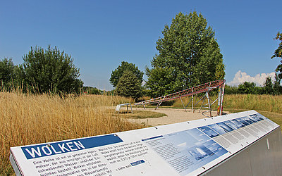 Vergrößerte Ansicht: Informationstafel mit Blick über eine Wiese mit Bäumen zum blauen Himmel
