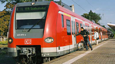 Red suburban train at a platform. One person with a bike pressing button to open doors.