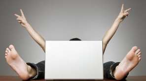 A man sits hidden behind a laptop. You can only see his arms and feet.