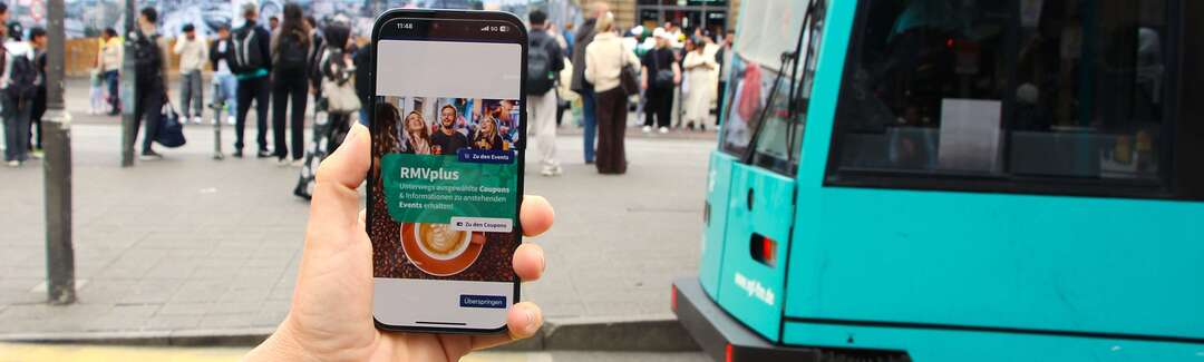 Hand mit Handy. Auf dem Display ist die RMVplus-App zu sehen
