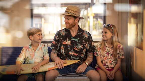 S-Bahn-Kampagne Urlaub Ein Mann mit Sonnenhut sitzt mit Kindern und Surfboard in einer Bahn