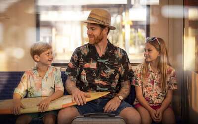 Ein Mann mit Sonnenhut sitzt mit Kindern und Surfboard in einer Bahn