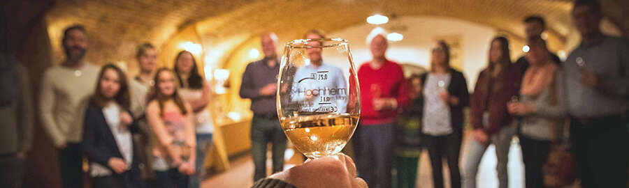 Hand hält ein gefülltes Weinglas, dahinter eine Besuchergruppe in einem Gewölbe-Keller