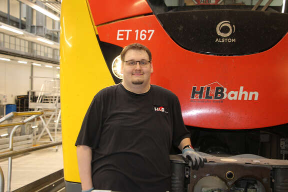 Junger Mann mit Brille steht vor einem schwarz-rot-gelben Zug. Neben ihm die Aufschrift "HLB Bahn"