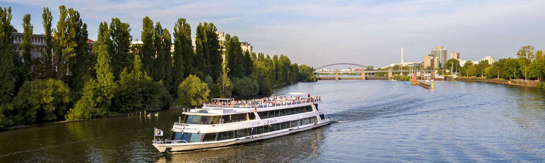 Ausflugsschiff auf dem Main vor baumbesäumten Ufern, im Hintergrund Schleuse, Brücke und Hochhäuser