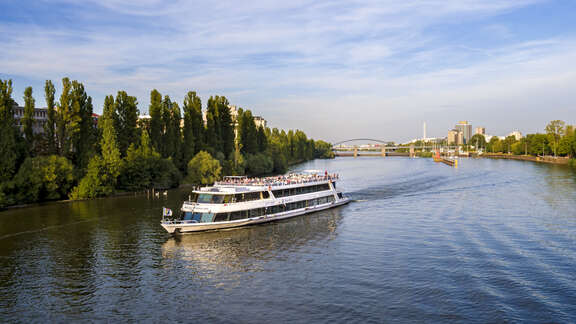 Ausflugsschiff auf dem Main vor baumbesäumten Ufern, im Hintergrund Schleuse, Brücke und Hochhäuser