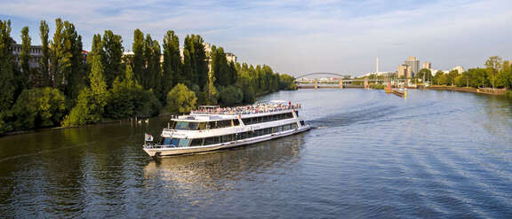 Ausflugsschiff auf dem Main vor baumbesäumten Ufern, im Hintergrund Schleuse, Brücke und Hochhäuser