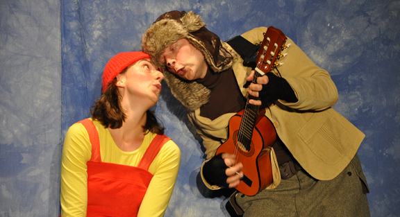 Zwei Menschen gucken sich an, die Frau mit Latzhose und Mütze, der Mann mit einer Gitarre in der Hand 
