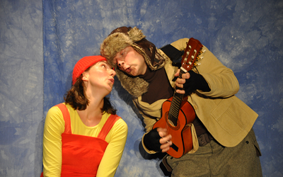 Zwei Menschen gucken sich an, die Frau mit Latzhose und Mütze, der Mann mit einer Gitarre in der Hand 