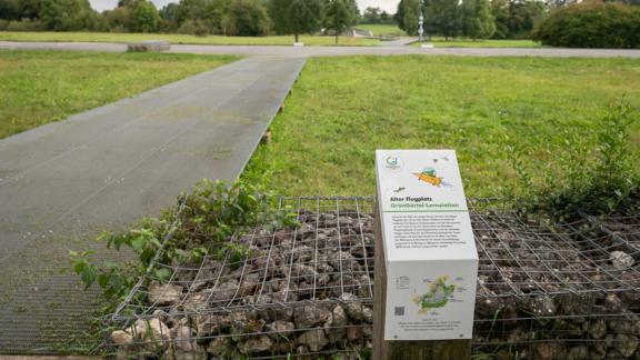info-'Tafel am Alten Flugplatz Bonames mit Grünfläche im Hintergrund.