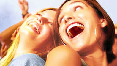 zwei junge Frauen lächeln sich fröhlich an, im Hintergrund erhobene Hände von feiernden Menschen