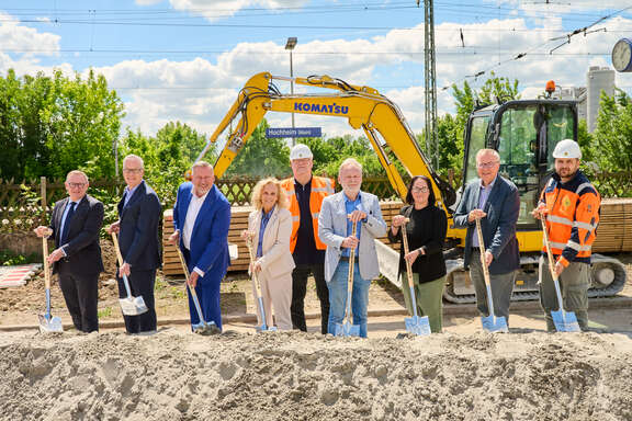 Neun Personen stehen nebeneinander stehen nebeneinander vor einem Sandhügel mit einer Schaufel in der Hand. Im Hintergrund das Stationsschild Hochheim