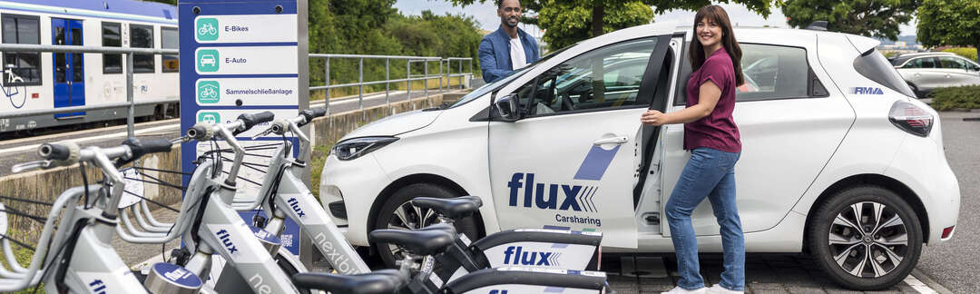 Flux Car- und Bikesharing An einer Bahnstation stehen Flux Car- und Bikesharingangebote. Ein Paar steigt in ein Auto ein.
