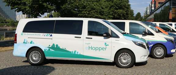 Ein weißer Van steht auf einem Platz unter Bäumen, Aufschrift: On-Demand-Shuttle, kvgOG Hopper