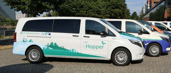 Ein weißer Van steht auf einem Platz unter Bäumen, Aufschrift: On-Demand-Shuttle, kvgOG Hopper