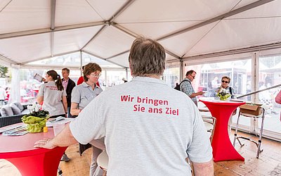 Ein Mann tragt ein Shirt mit der Rückenaufschrift "Wir bringen Sie ans Ziel". Im Hintergrund sieht man eine Gruppe von Menschen, die den MTV-Stand besuchen.