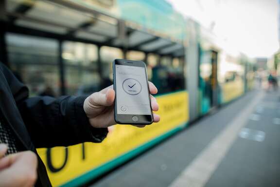 Eine Hand hält ein Smartphone, im Display steht: Fahrt aktiv; im Hintergrund: Straßenbahn und Haltestellenbereich