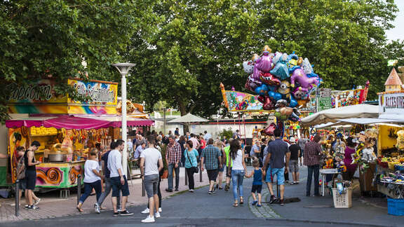Viele Menschen flanieren bei sonnigem Wetter an den Schaubuden vorbei. Dazwischen läuft ein Luftballonverkäufer.