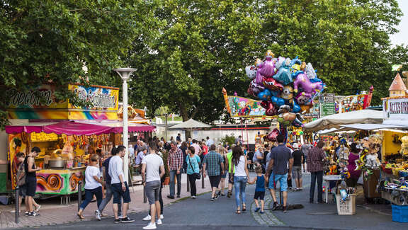 Viele Menschen flanieren bei sonnigem Wetter an den Schaubuden vorbei. Dazwischen läuft ein Luftballonverkäufer.