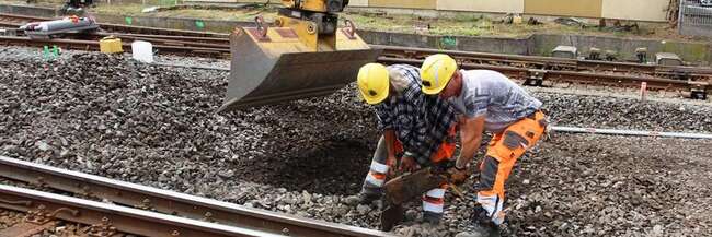 Zwei Baurbeiter bücken sich neben dem Schaufelkopf eines Baggers