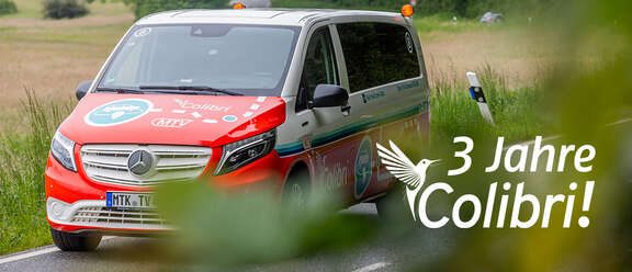 Ein rot-weißes Colibri-Fahrzeug mit Mercedes-Logo fährt auf einer Landstraße. Auf dem Fahrzeug sind bunte Grafiken und das Colibri-Logo zu sehen. Im Vordergrund sind Blätter unscharf, im Hintergrund eine Wiese. Auf dem Bild steht der Text: "3 Jahre Colibri!"