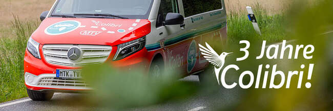 Ein rot-weißes Colibri-Fahrzeug mit Mercedes-Logo fährt auf einer Landstraße. Auf dem Fahrzeug sind bunte Grafiken und das Colibri-Logo zu sehen. Im Vordergrund sind Blätter unscharf, im Hintergrund eine Wiese. Auf dem Bild steht der Text: "3 Jahre Colibri!"
