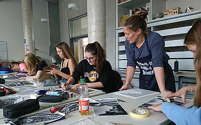 Vergrößerte Ansicht: ehrerin schaut Kunstschülern, die an einem langen Tisch arbeiten, beim Arbeiten zu