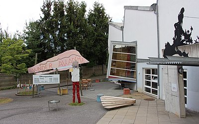 Vergrößerte Ansicht: Asymmetrisches Gebäude , bemaltes Schild mit Aufschrift Kinderakademie, Schild mit Öffnungszeiten