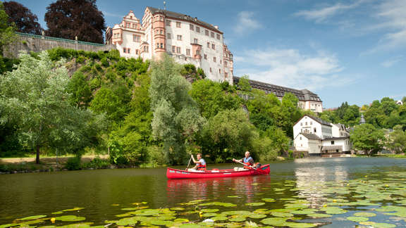 Zwei Menschen paddeln in einem roten Kanu über einen mit Seerosen bedeckten Fluss