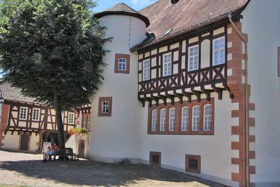 Vergrößerte Ansicht: Fachwerk-Haus mit seitlichem Rund-Turm, davor ein großer Baum mit Rund-Bank