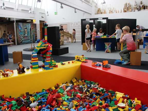 Vergrößerte Ansicht: riesige Spielkiste mit LEGO-duplo-Steinen, im Hintergrund: Kinder an Spieltischen in großer Halle