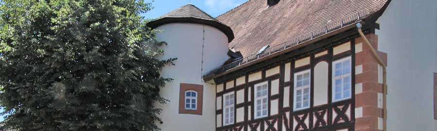 Fachwerk-Haus mit seitlichem Rund-Turm, davor ein großer Baum mit Rund-Bank