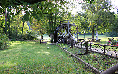 Vergrößerte Ansicht: Großes Wasserrad aus Holz, umgeben von Wiese und Bäumen 