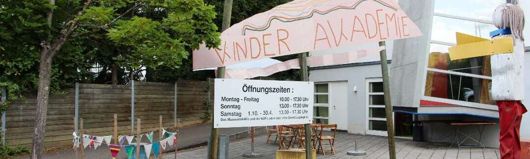 asymmetrisches Gebäude, bemaltes Schild mit Aufschrift Kinder Akademie, Schild mit Öffnungszeiten