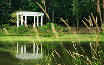 Vergrößerte Ansicht: Im Vordergrund ein Teich/See, im Hintergrund grüne Wiese mit einer weißen Terrasse 