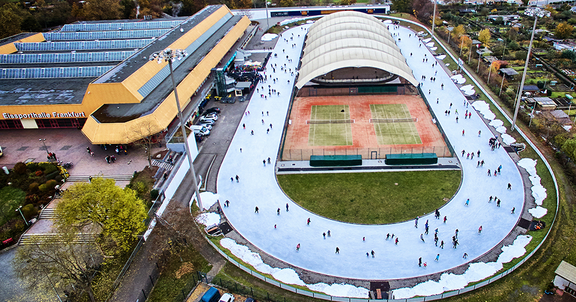 Eine ovale Eisfläche, in der Mitte eine überdachte Fläche und daneben zwei Tennisplätze
