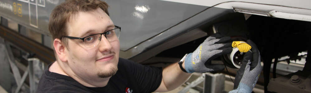 Ein junger Mann im schwarzen Shirt steht seitlich an einem grauen Zug. Mit zwei Handschuhen greift an ein schwarz-weiße Befestigung.