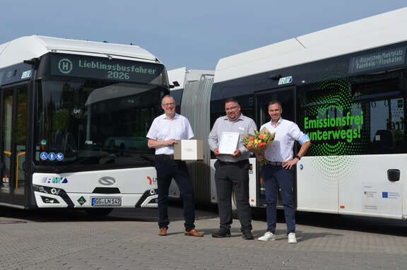 Drei Männer stehen vor zwei Bussen und halten zusammen eine Urkunde in den Händen