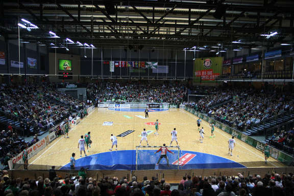 Two handball teams play on the field in front of an audience