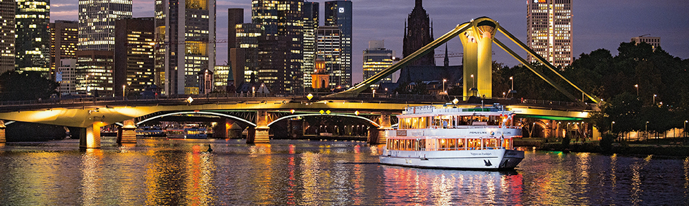 Auf dem Main ist ein Schiff vor einer Brücke. Im Hintergrund Hochhäuser mit Lichtern