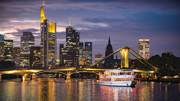 Auf dem Main ist ein Schiff vor einer Brücke. Im Hintergrund Hochhäuser mit Lichtern