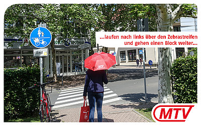 Eine Frau mit rotem MTV-Regenschirm steht vor einem Zebrastreifen. Die Globus Apotheke befindet sich im Hintergrund: ...laufen nach links Über den Zebrastreifen und gehen einen Block weiter...