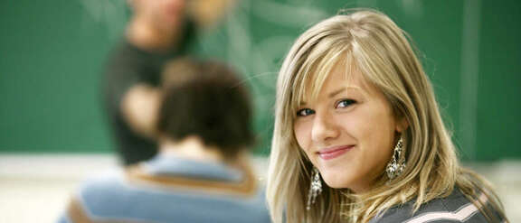 A girl in a classroom looking in the camera, behind her a teacher showing things on a blackboard