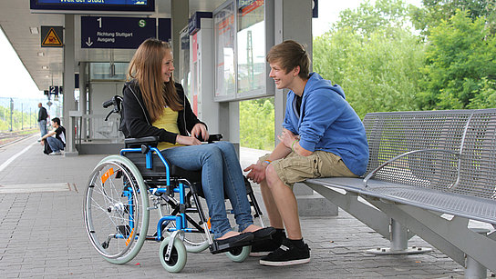 Junge Frau im Rollstuhl am Bahngleis mit einem männlichen Begleiter