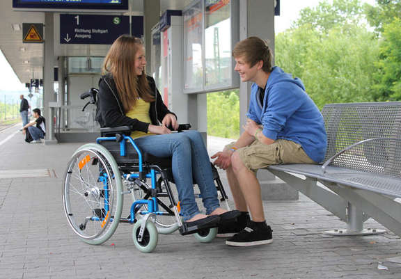 Junges Mädchen im Rollstuhl am Bahngleis mit einem männlichen Begleiter