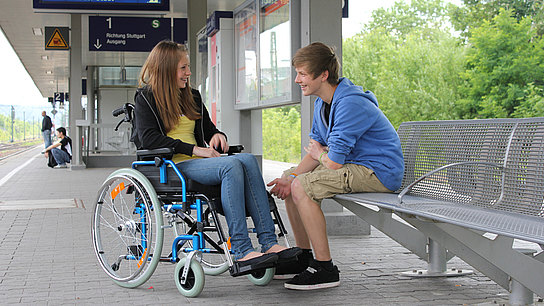 Junge Frau im Rollstuhl am Bahngleis mit einem männlichen Begleiter