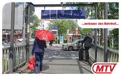 Eine Frau mit rotem MTV-Regenschirm verlässt den Bahnhof. Ein Anruf-Sammelt-Taxi steht im Hintergrund: ...verlassen den Bahnhof...
