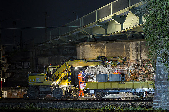 Schienenbagger und Anhänger mit Bauelementen unter Brücke Schienenbagger und Anhänger mit Bauelementen unter einer beleuchteten Brücke