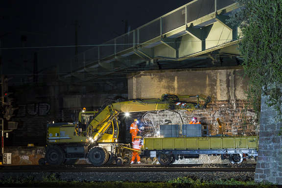 Schienenbagger und Anhänger mit Bauelementen unter Brücke Schienenbagger und Anhänger mit Bauelementen unter einer beleuchteten Brücke